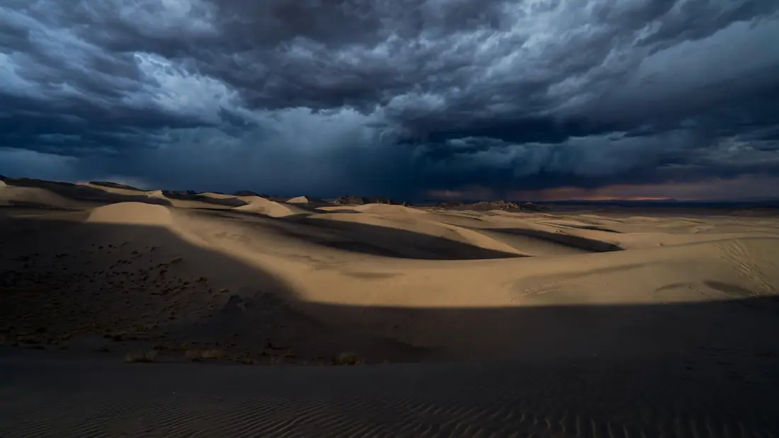 Dark heavy storm clouds gathering over a desert at dusk, deep shadows spreading across empty dunes