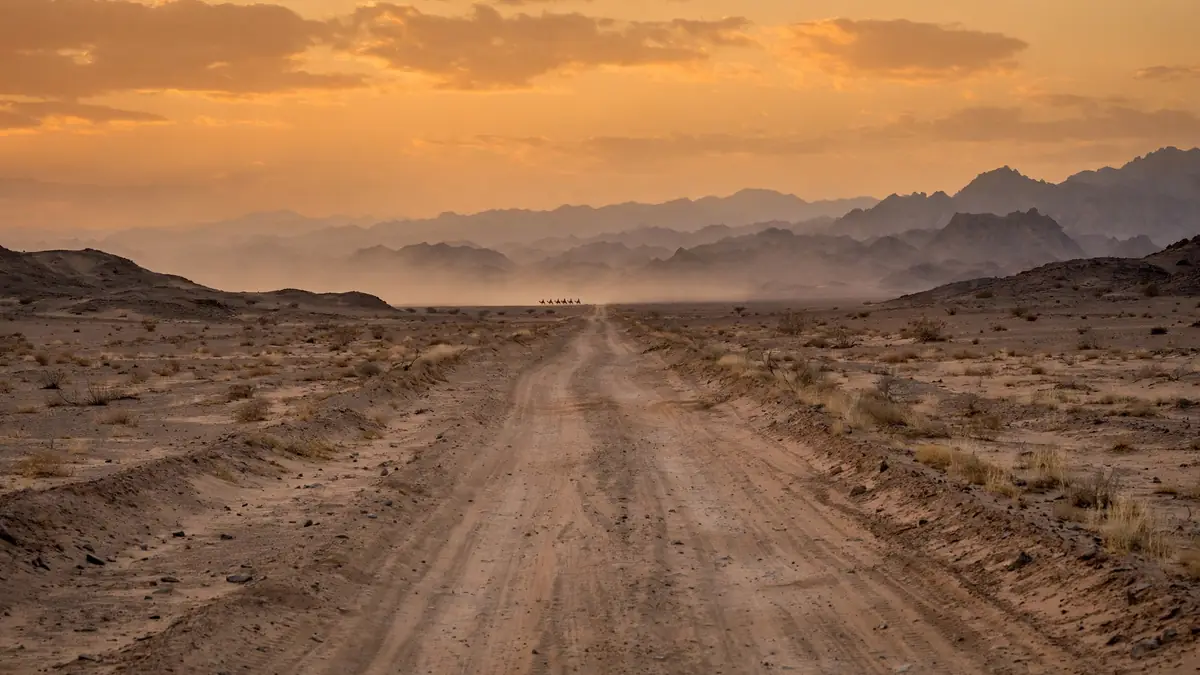A dusty caravan road stretching east toward distant mountains at dusk, the direction of his emergence