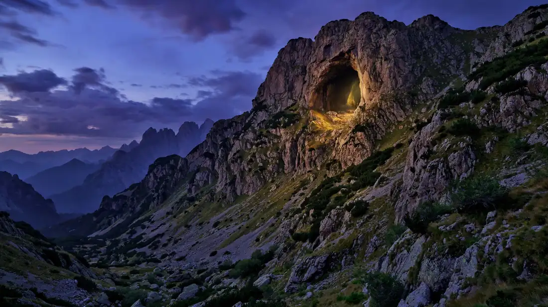 A dark cave mouth high in a rugged mountain at dusk, a faint light glowing from within